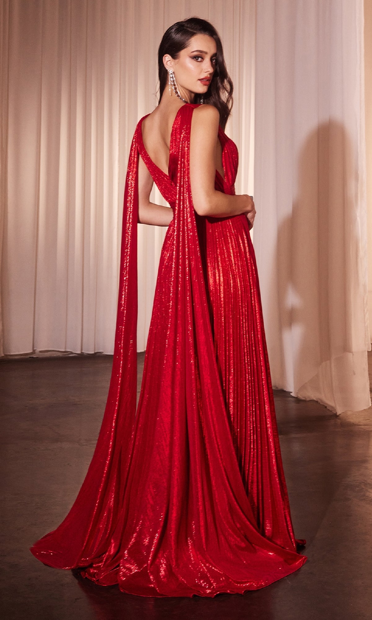 A woman wearing the Ladivine CD802 Long Prom Dress, featuring a shimmering red A-line pleated skirt and draped back, stands indoors in front of white curtains.